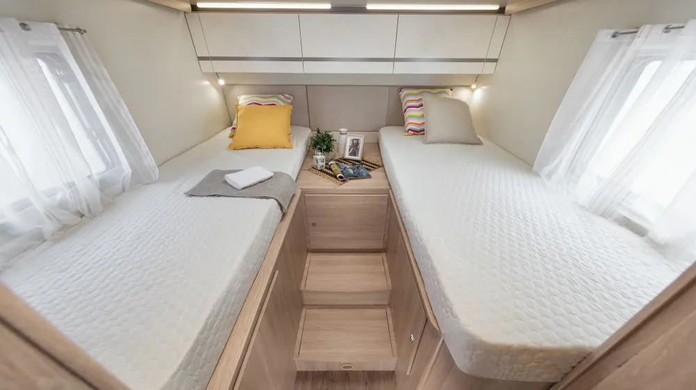 Dos camas simples con sabanas blancas y cojines amarrillo y blanco. Ventanas y cortinas blancas. Imagen por dentro de autocaravana Giotti Siena 395